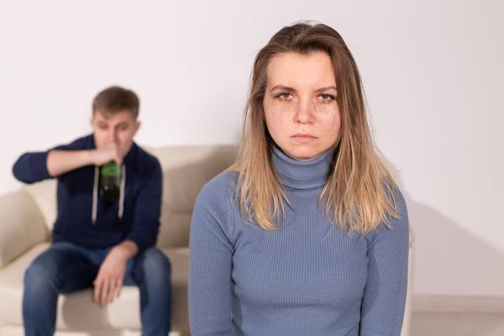 My Partner Abuses Me When I'm Already Hurting 10 Angry wife with husband in background drinking