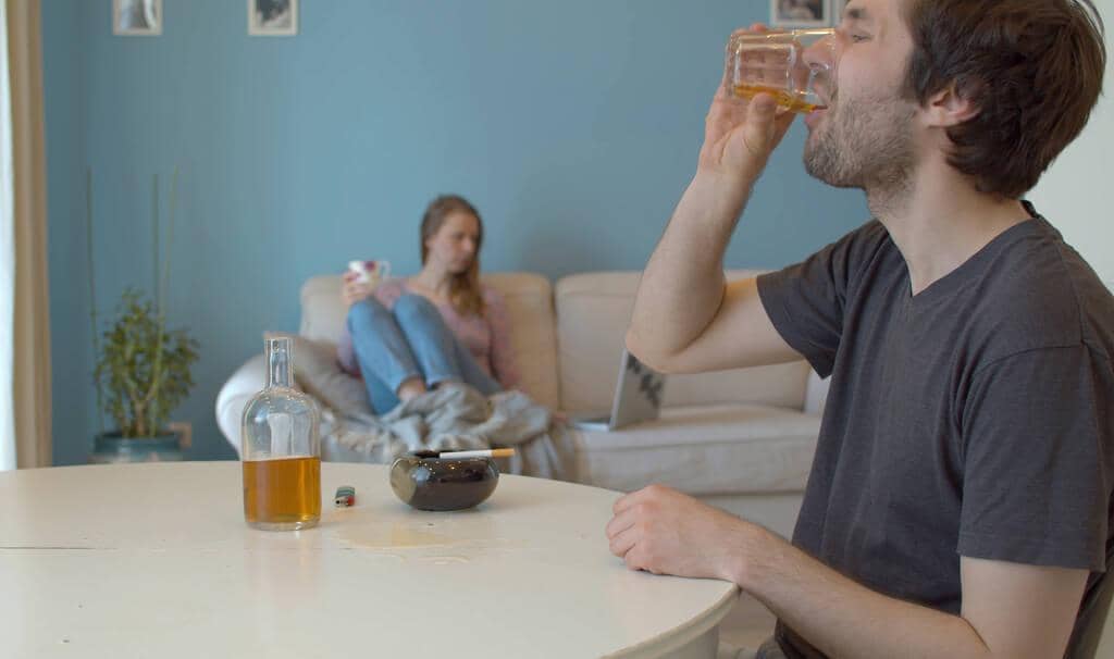 With One Partner Still Partying Can Your Relationship Thrive? 2 man drinking whiskey background defocus wife sits couch looks
