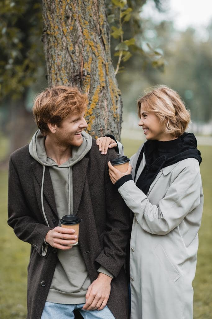 2025’s Most Toxic Dating Trends: Behaviors You Need to Avoid 8 happy blonde woman holding paper cup laughing boyfriend tree trunk