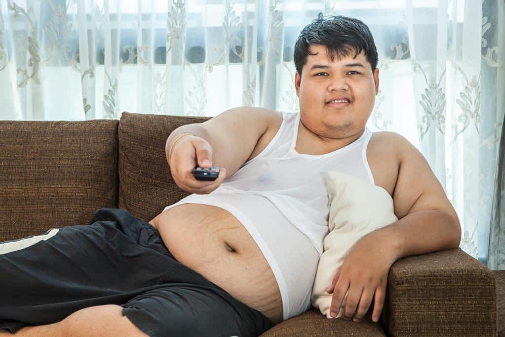 Overweight asian guy sitting on the couch with remote in hand trying to watch some TV