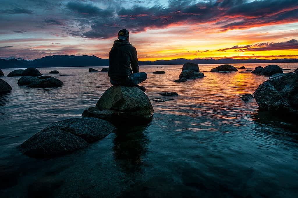 man sitting on boulders at the ocean watching a sunset