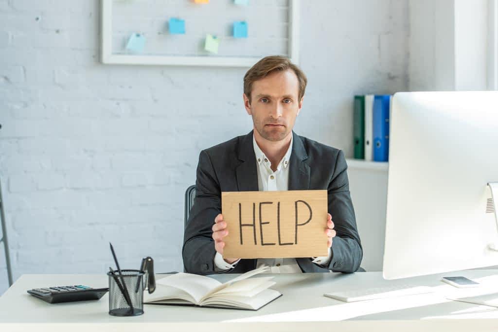 sad businessman looking camera holding cardboard help lettering while sitting