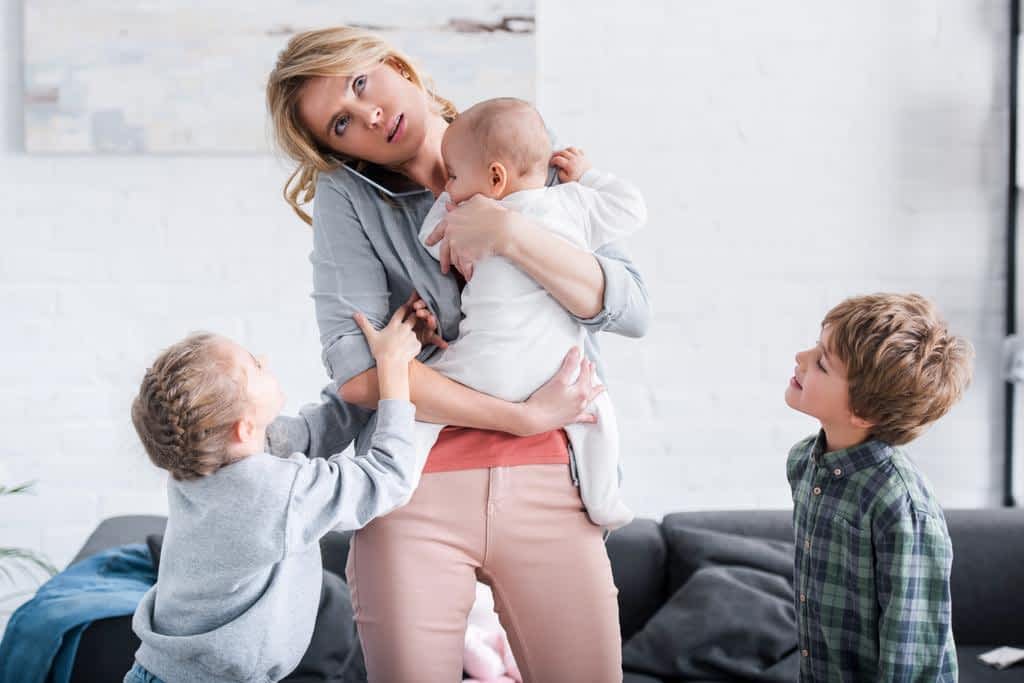 tired mother talking smartphone holding infant child while siblings standing