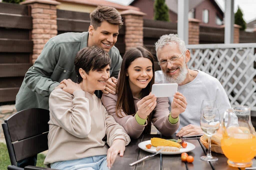 smiling teen girl using smartphone middle aged parents brother barbeque