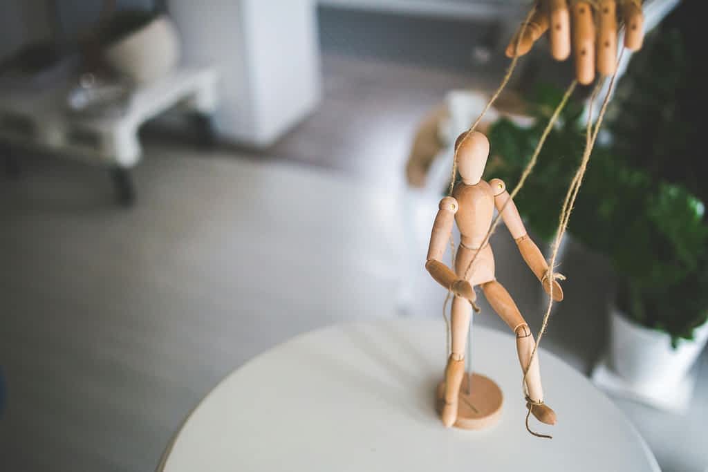 hands controlling a wooden puppet depicting control