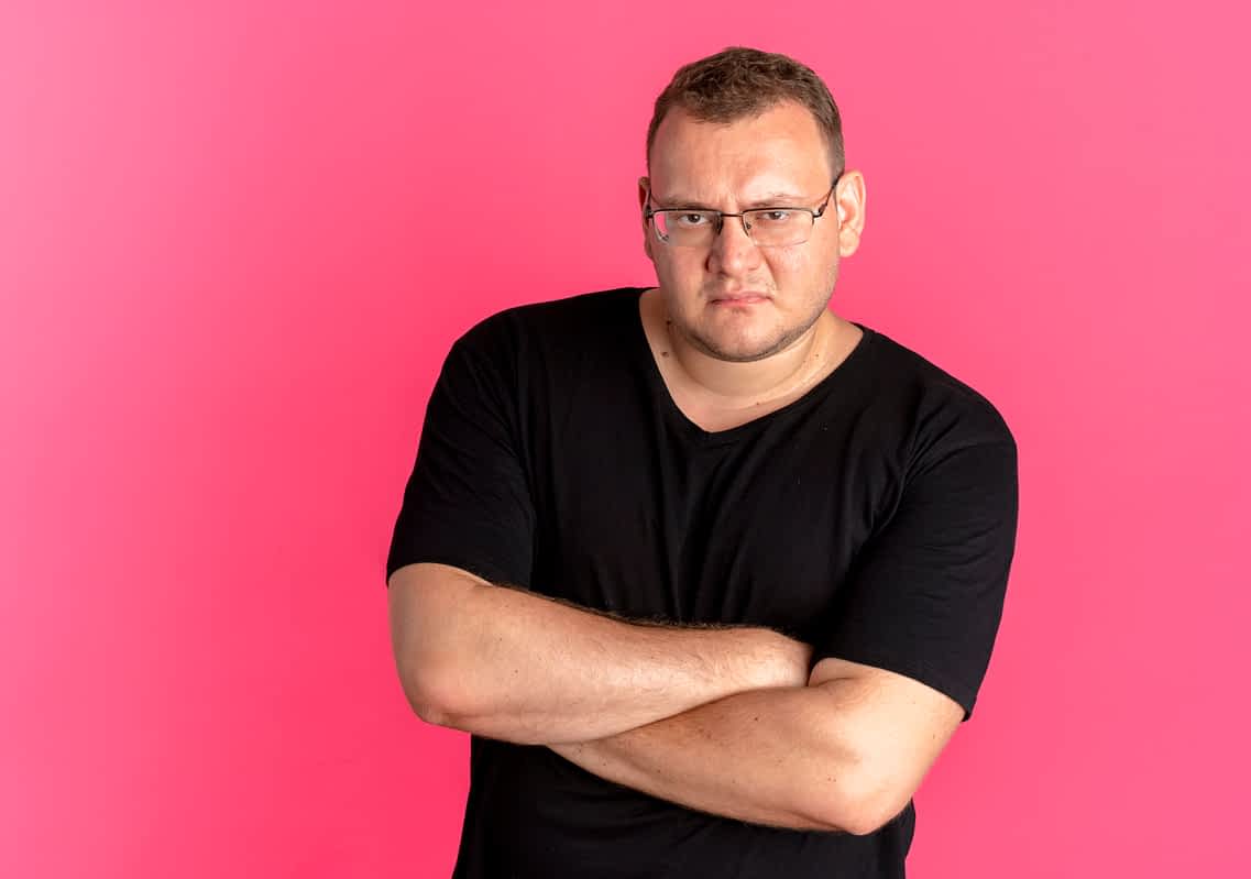 resentful overweight man in glasses wearing black t-shirt looking at camera with frowning face with crossed arms standing over pink background