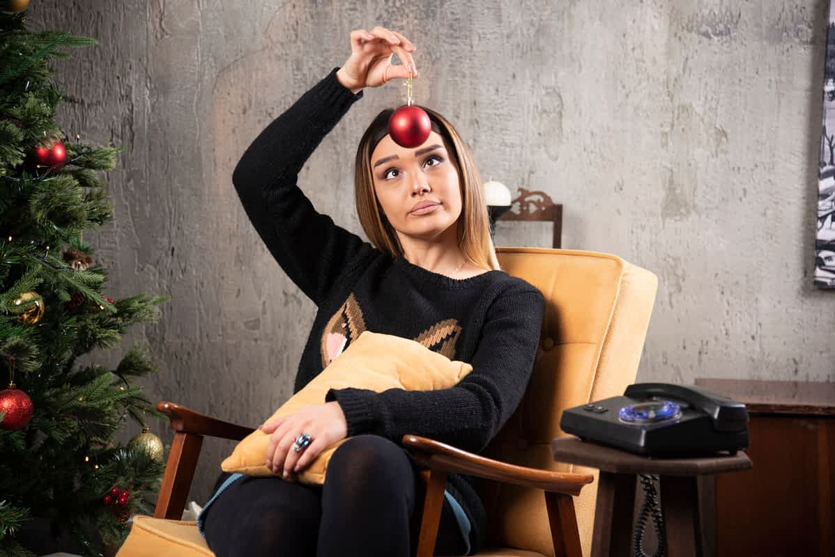 tired woman looking at ball cross-eyed next to Christmas tree.