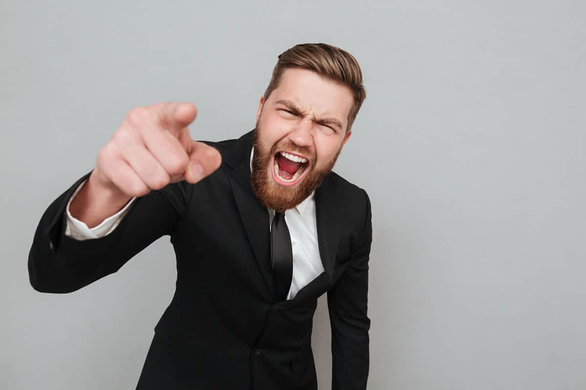 Angry furious businessman in suit shouting and pointing finger at camera isolated over gray background