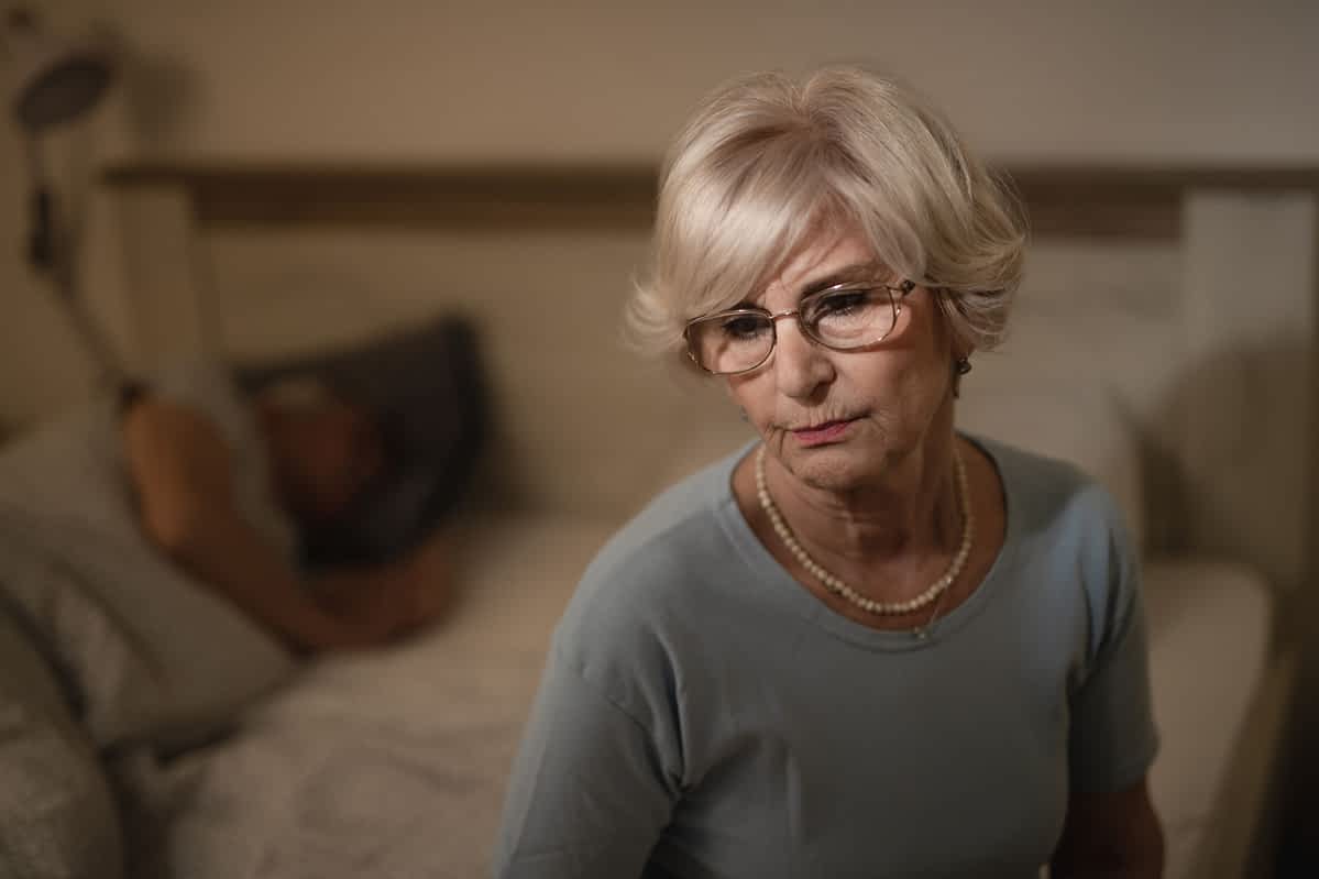 Thoughtful mature woman sitting in bedroom in the evening while her husband is sleeping in the background.