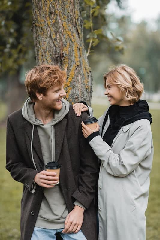 happy blonde woman holding paper cup laughing boyfriend tree trunk