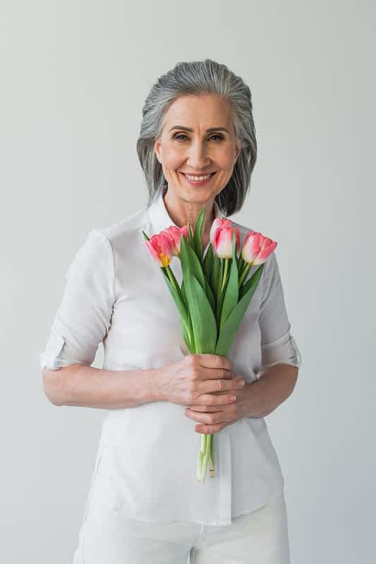 9 Reasons Men Prefer Younger Women: Keeping Your Confidence Strong 4 positive mature woman white shirt holding tulips isolated grey