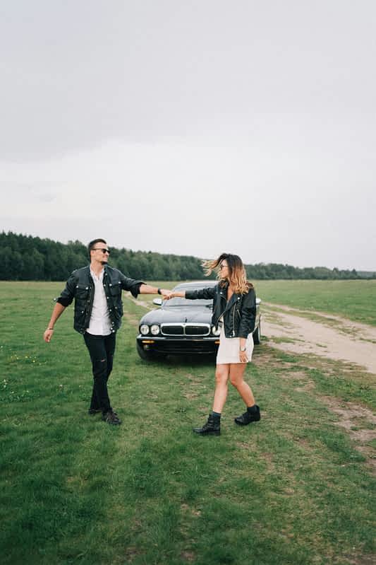 Man and woman dance in a green field in front of a black car