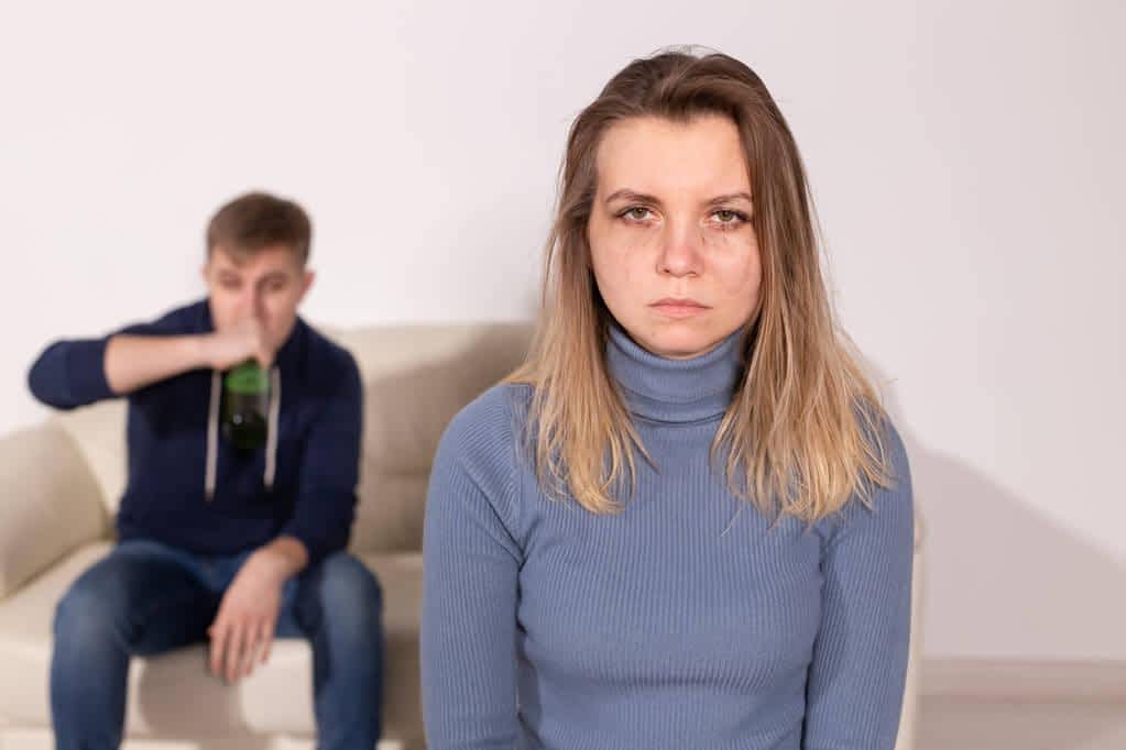 Angry wife with husband in background drinking