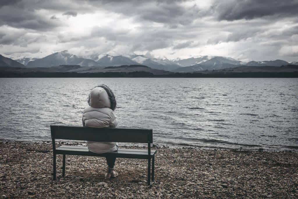 woman jacket sitting bench shore lake dark cloudy sky mountains 1
