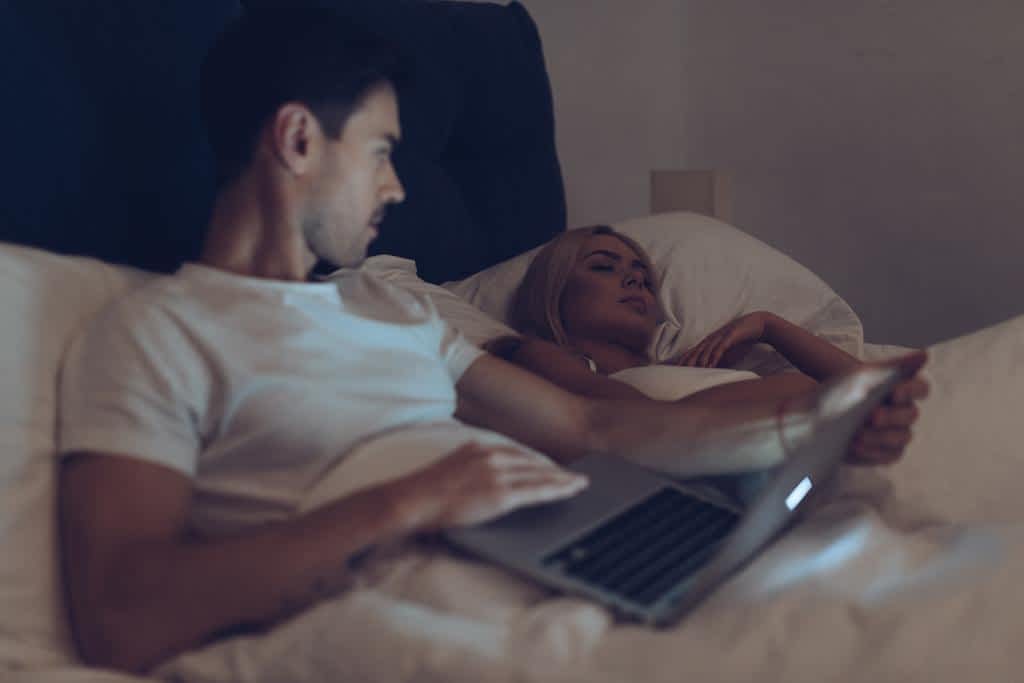 young man using laptop looking girlfriend sleeping bed night
