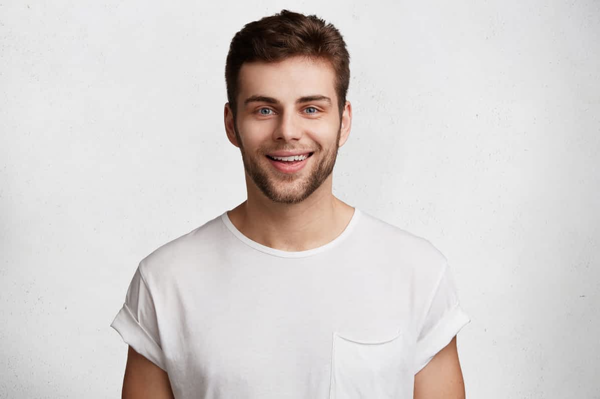 A bare minimum man in a white t-shirt smiling against a gray background