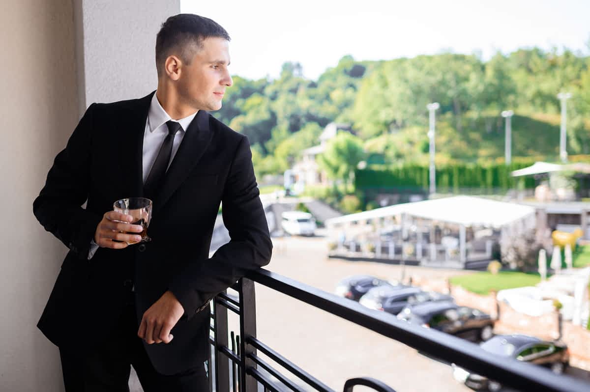 Side view of handsome groom in wedding black suit, holding glass with drink while leaning on balcony and looking away on nature landscape