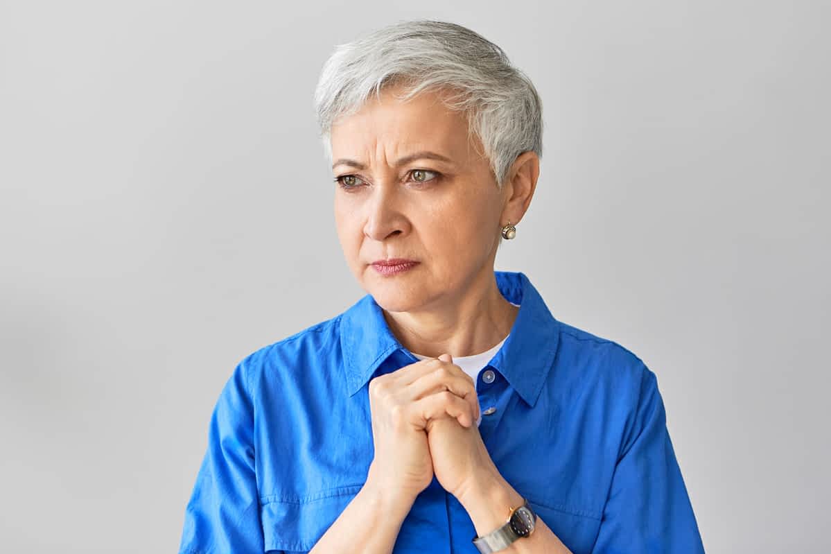Portrait of serious frowning middle aged mature European woman with gray pixie hair expressing nervousness, holding clasped hands on her chest, being impatient, waiting.